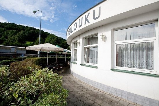 Zaoblená bílá fasáda restaurace V Oblouku s nápisem na římse, vedle ní terasa se slunečníky